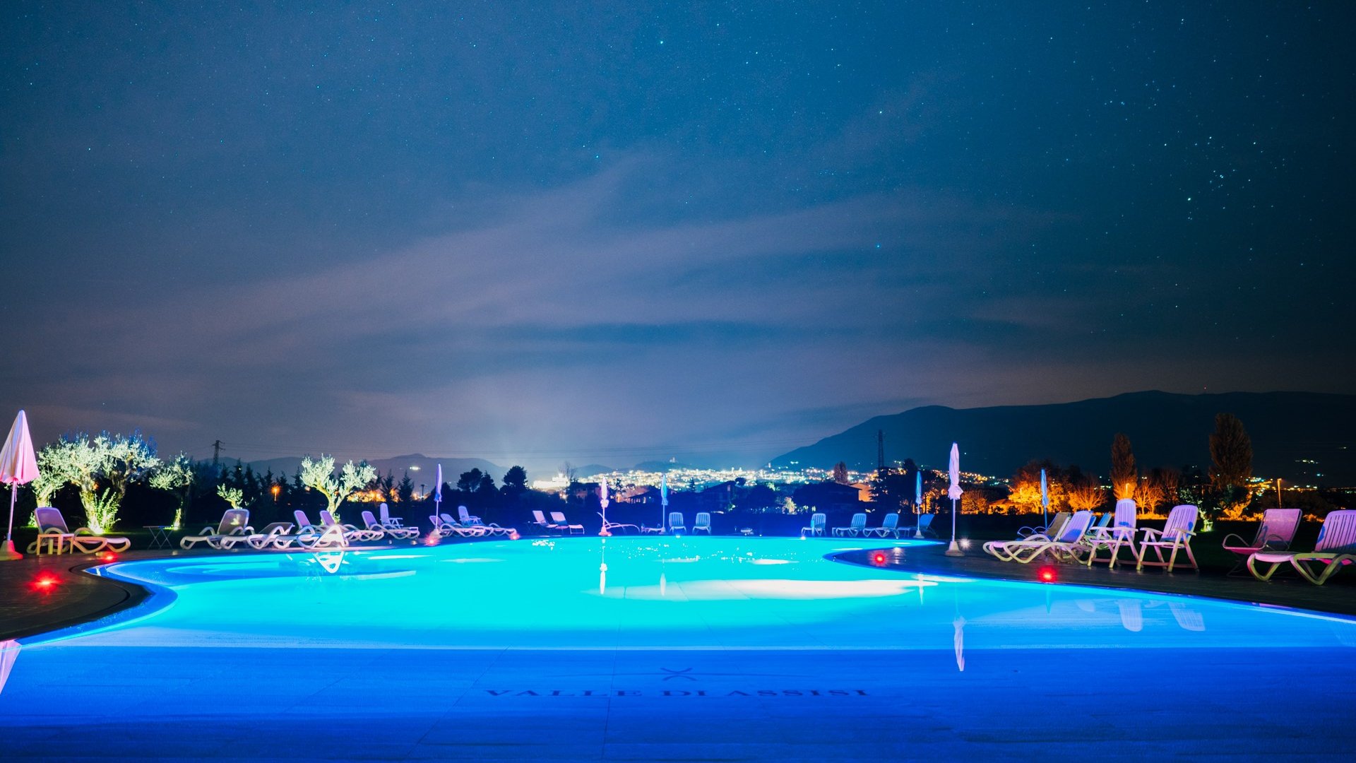 Serata di Astronomia Umbria Assisi con Cena sotto le Stelle Piscina illuminata di notte con lettini e ombrelloni e vista sulle luci della città
