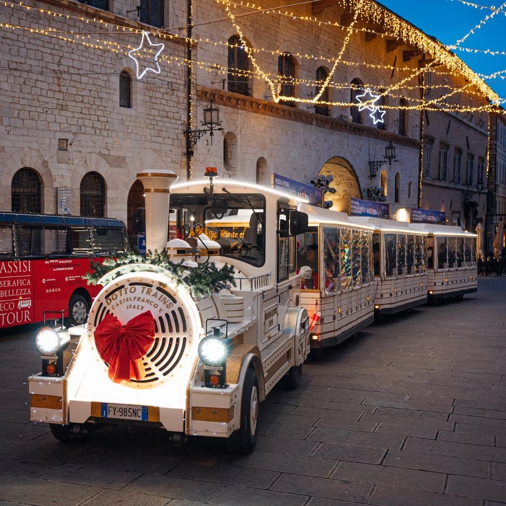 Natale e Inizio Anno a Valle di Assisi 2025 Trenino turistico bianco decorato con luci natalizie in una piazza di sera