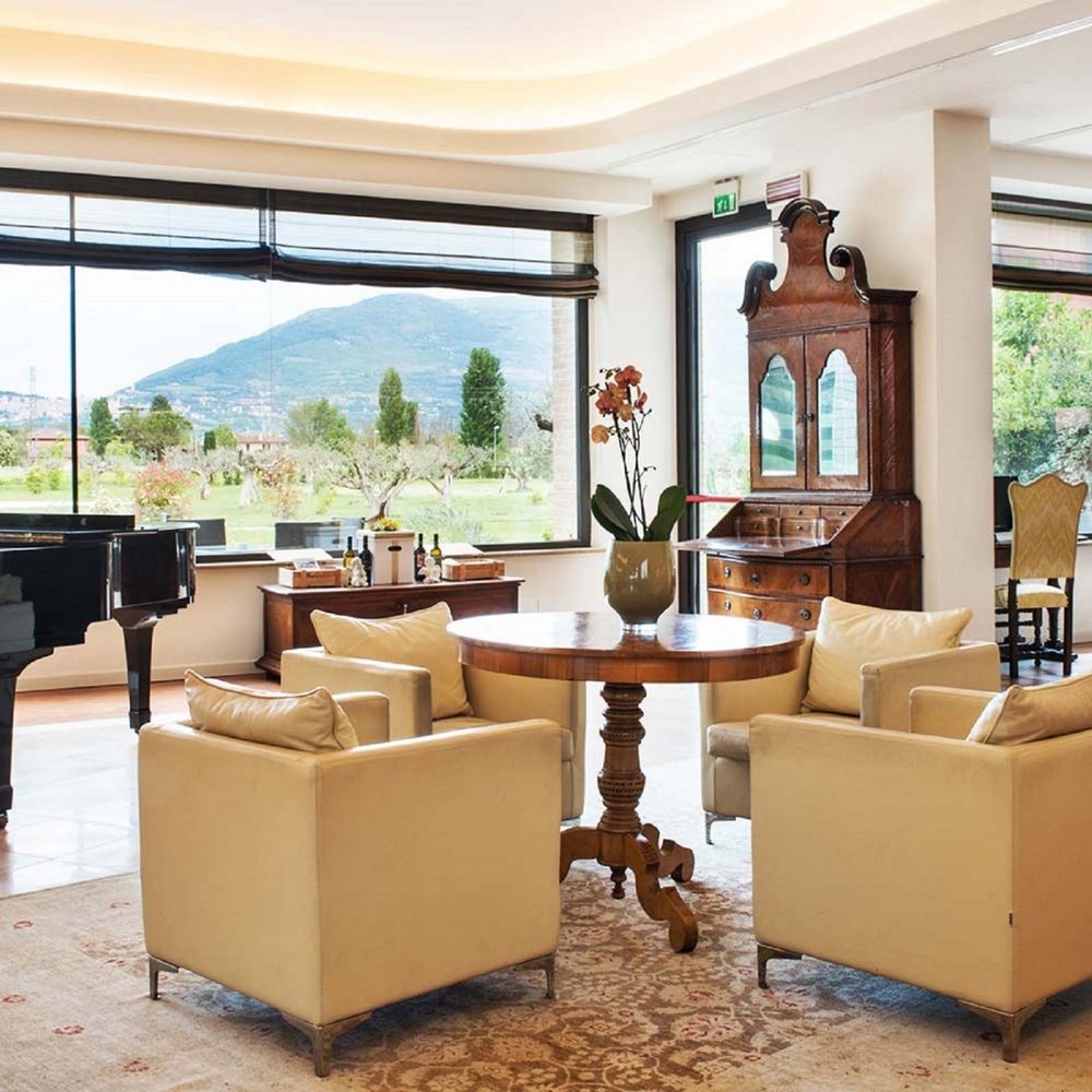 Un hotel per famiglie, in Umbria, con una marcia in più Salotto elegante con poltrone beige, tavolo rotondo, pianoforte e vista panoramica sulle montagne