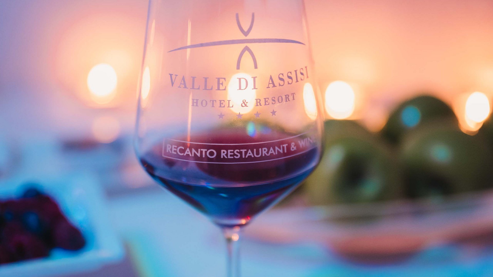 Gift Voucher Idee Regalo Festa della Mamma e anniversari dalla SPA benessere ai massaggi con cena. Calice di vino rosso con logo Valle di Assisi Hotel e Resort e candele sullo sfondo