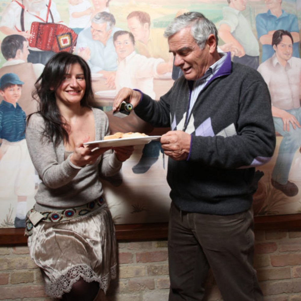 Natale e Inizio Anno a Valle di Assisi 2025 Donna che sorride con piatto mentre uomo versa salsa in un locale con muro dipinto
