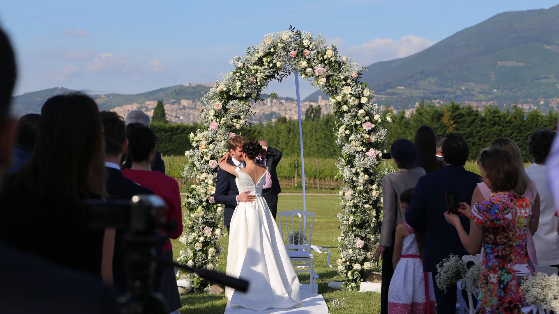 Valle di Assisi: un resort per matrimoni ad Assisi Sposi che si baciano sotto un arco di fiori durante una cerimonia all'aperto